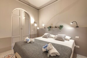 Ein ordentlich bezogenes Bett mit grauer Decke und Handtüchern, eine Nachttischlampe und ein an der Wand befestigtes Regal mit Dekoration.