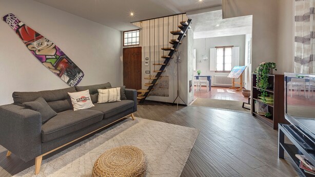 Un salon moderne avec un canapé gris, un grand tableau abstrait, un escalier et un coin repas visible à travers une porte ouverte.