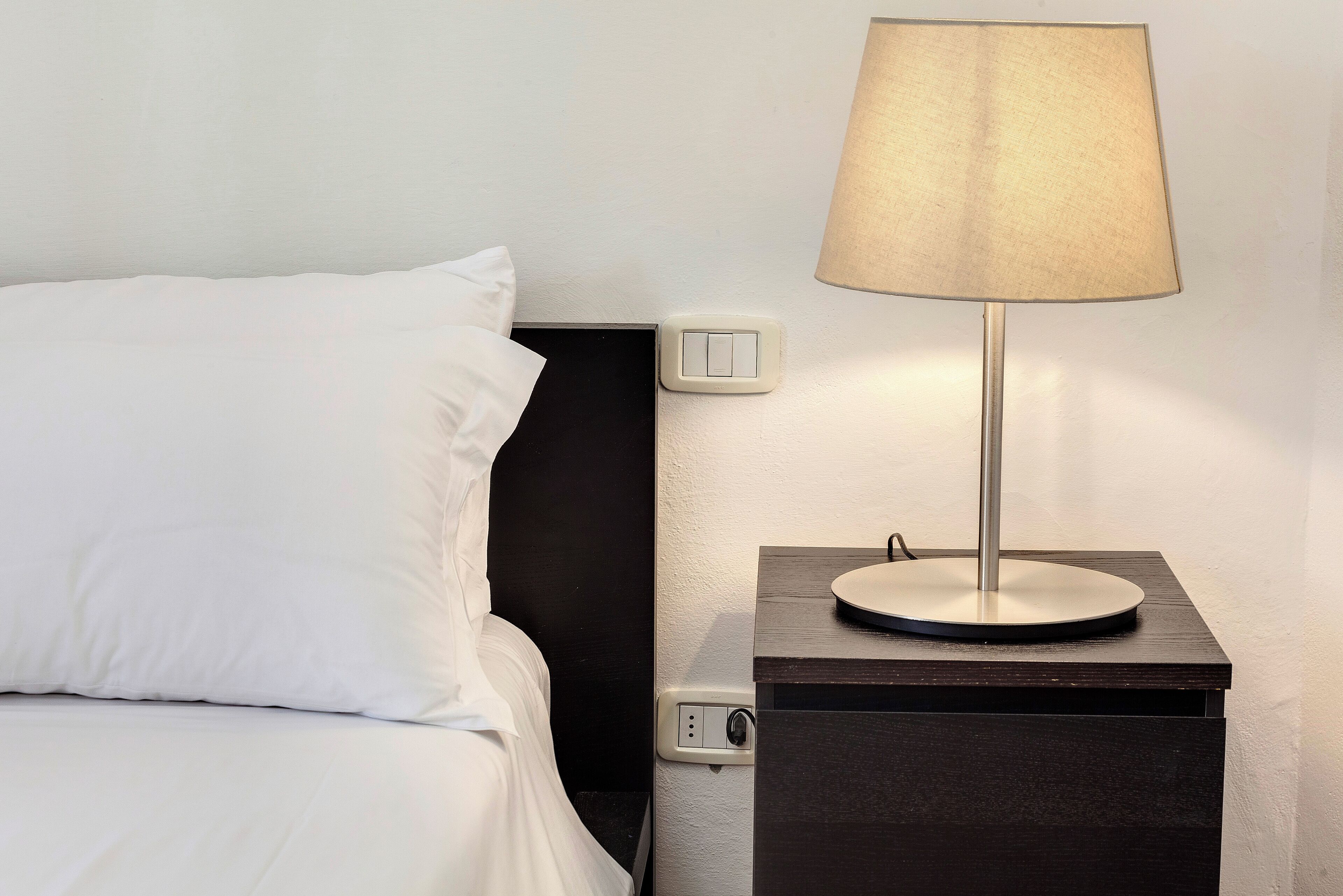 Un lit bien fait, avec du linge de lit blanc, une table de chevet avec une lampe et une prise électrique sur le mur.