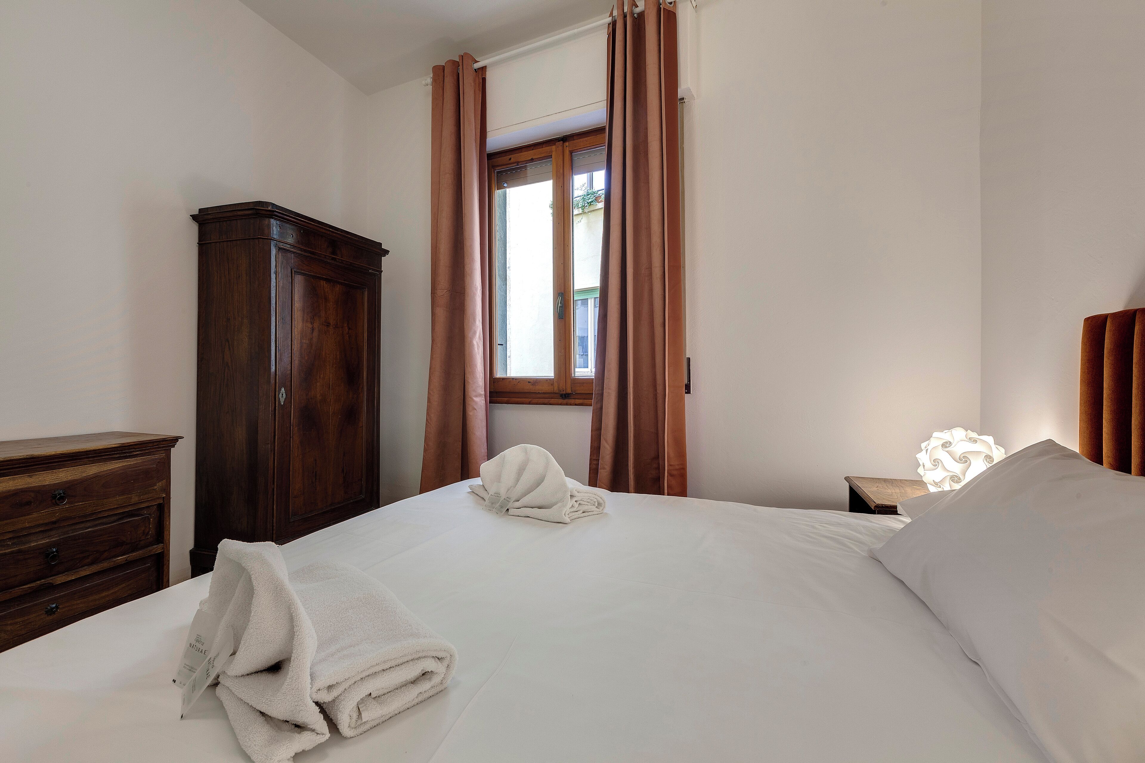 A neatly made bed with a white comforter and pillows, a wooden dresser, and a window with curtains.