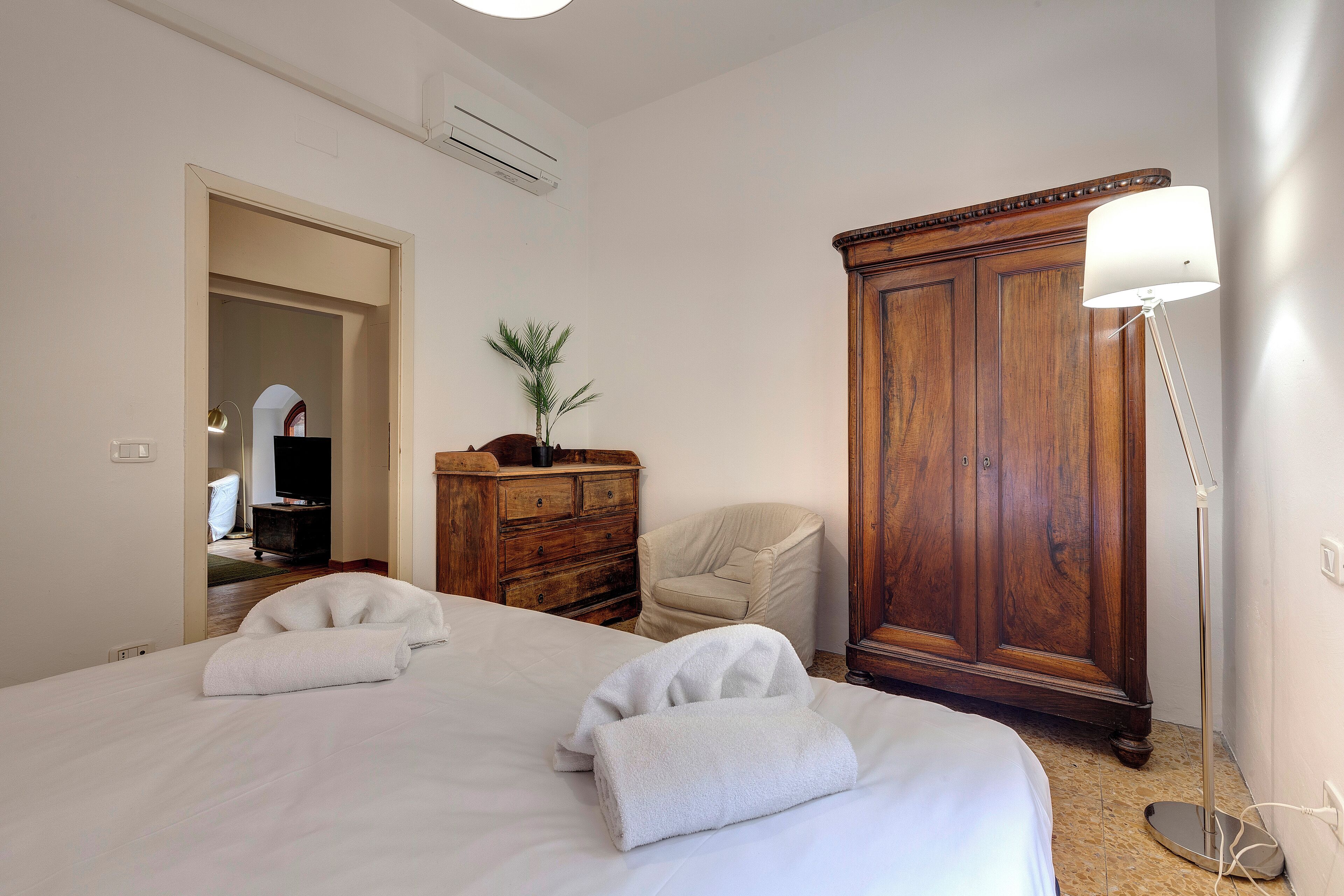 A hotel room with a large bed, a wooden wardrobe, a desk with a lamp, and a chair.