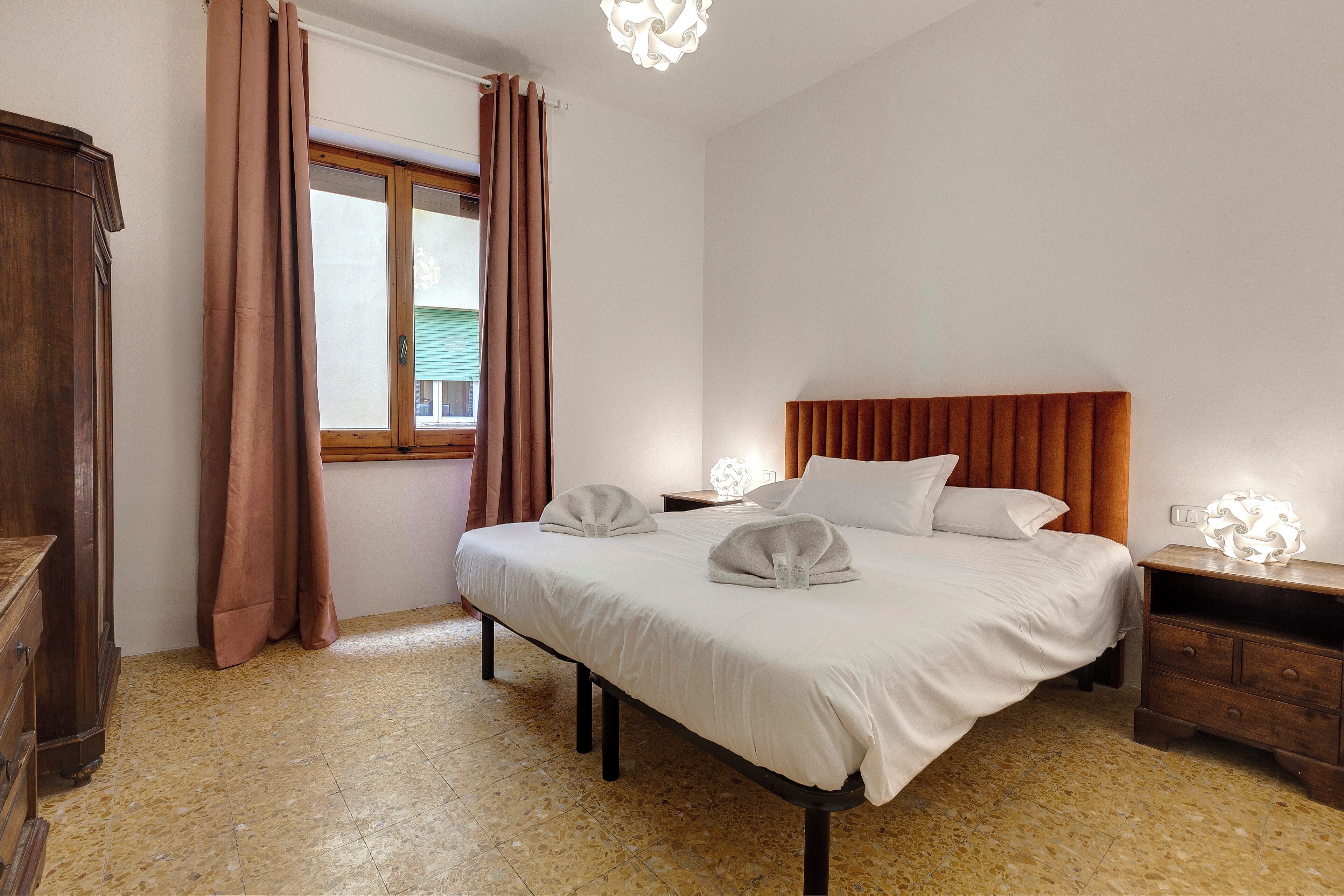 Un lit bien fait, avec des oreillers et une couverture, une table de chevet en bois avec une lampe, et une fenêtre avec des rideaux.