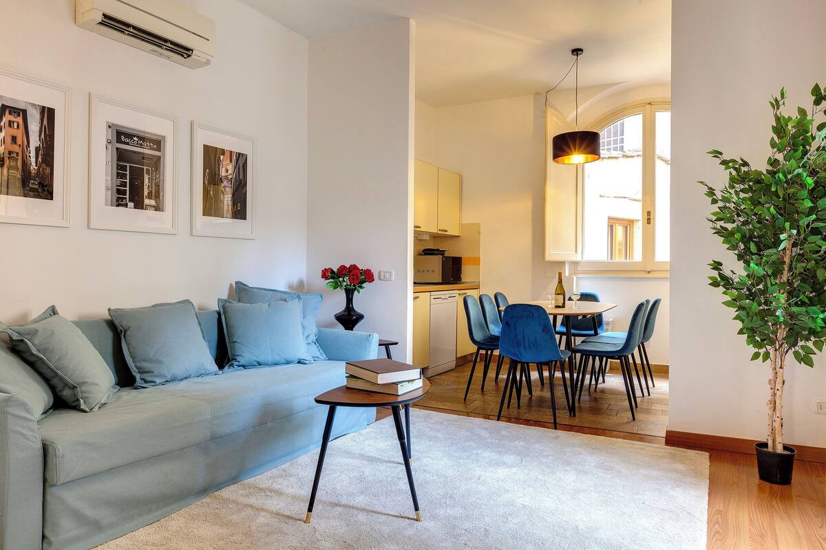 A living room with a sofa, a coffee table, and a dining area with chairs and a table. There is a potted plant and framed pictures on the wall.