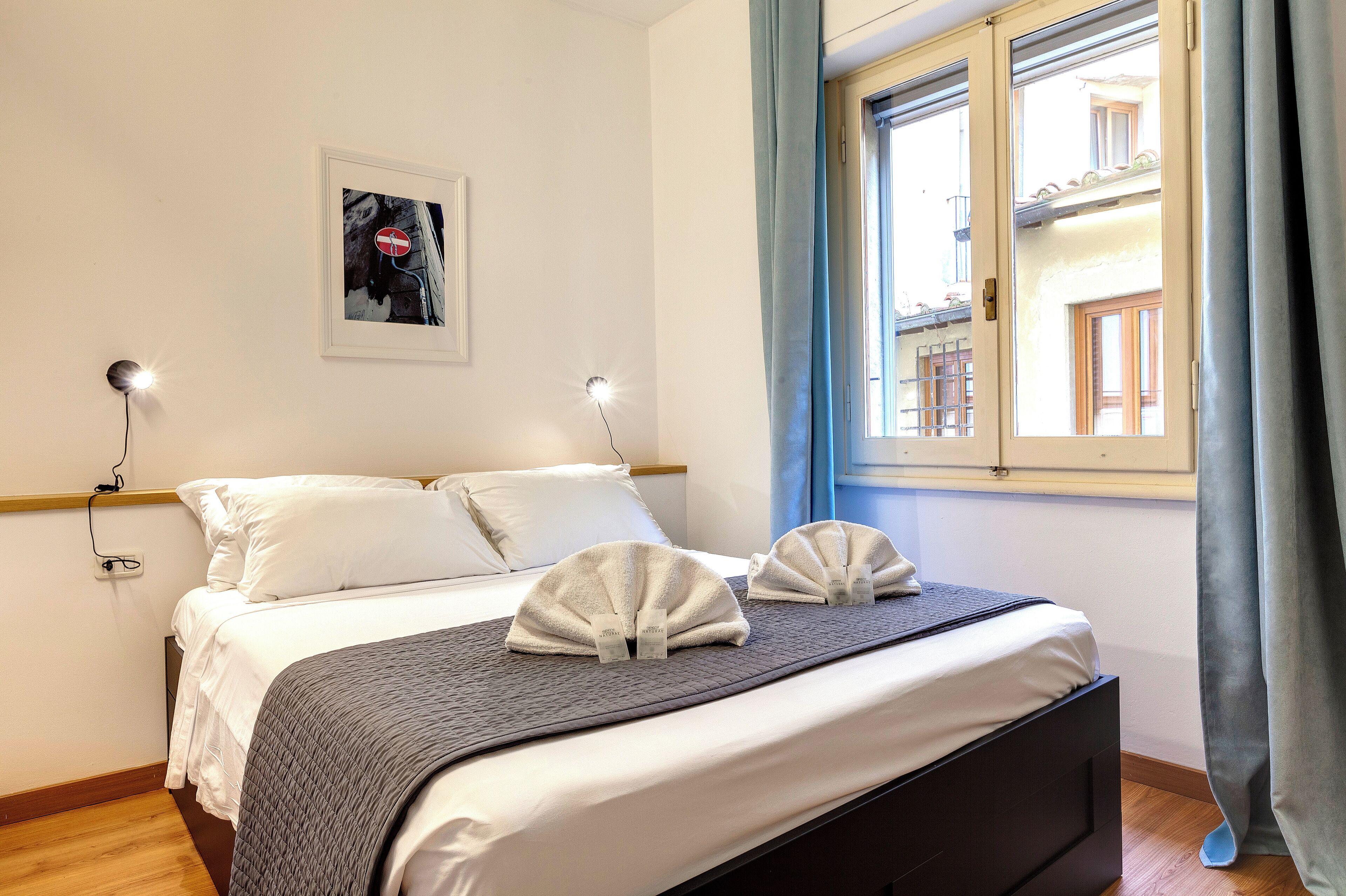 Ein ordentlich bezogenes Bett mit weißen Leinen und zwei Handtüchern, eine Nachttischlampe, ein gerahmtes Bild an der Wand und ein Fenster mit Vorhängen.