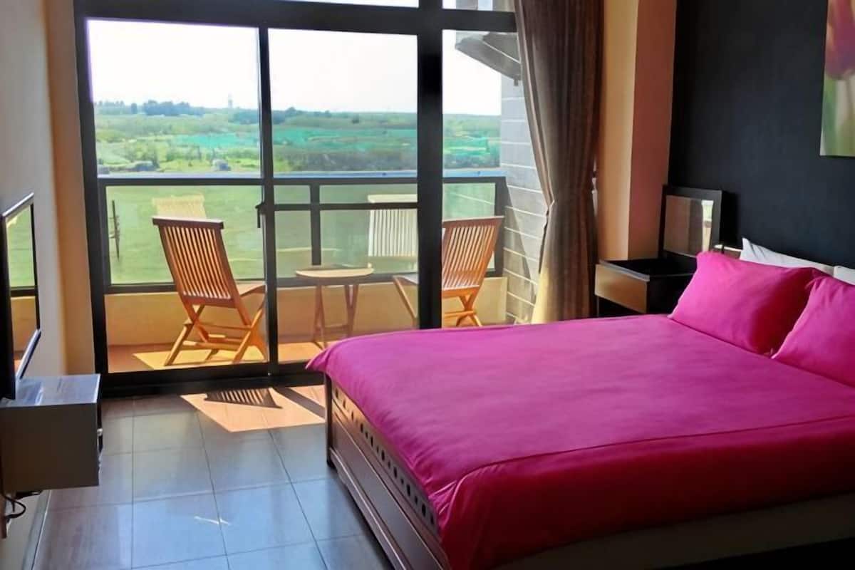 A bedroom with a large bed, a balcony with chairs, and a view of the countryside.