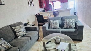 A living room with two grey sofas, a coffee table with decorative items, and a kitchen in the background. - 3-bedroom house in brilliant Potters Village with AC (Potters Village)