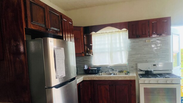 A kitchen with dark wooden cabinets, a stainless steel refrigerator, a white stove, and a window with a white curtain. - 3-bedroom house in brilliant Potters Village with AC (Potters Village)