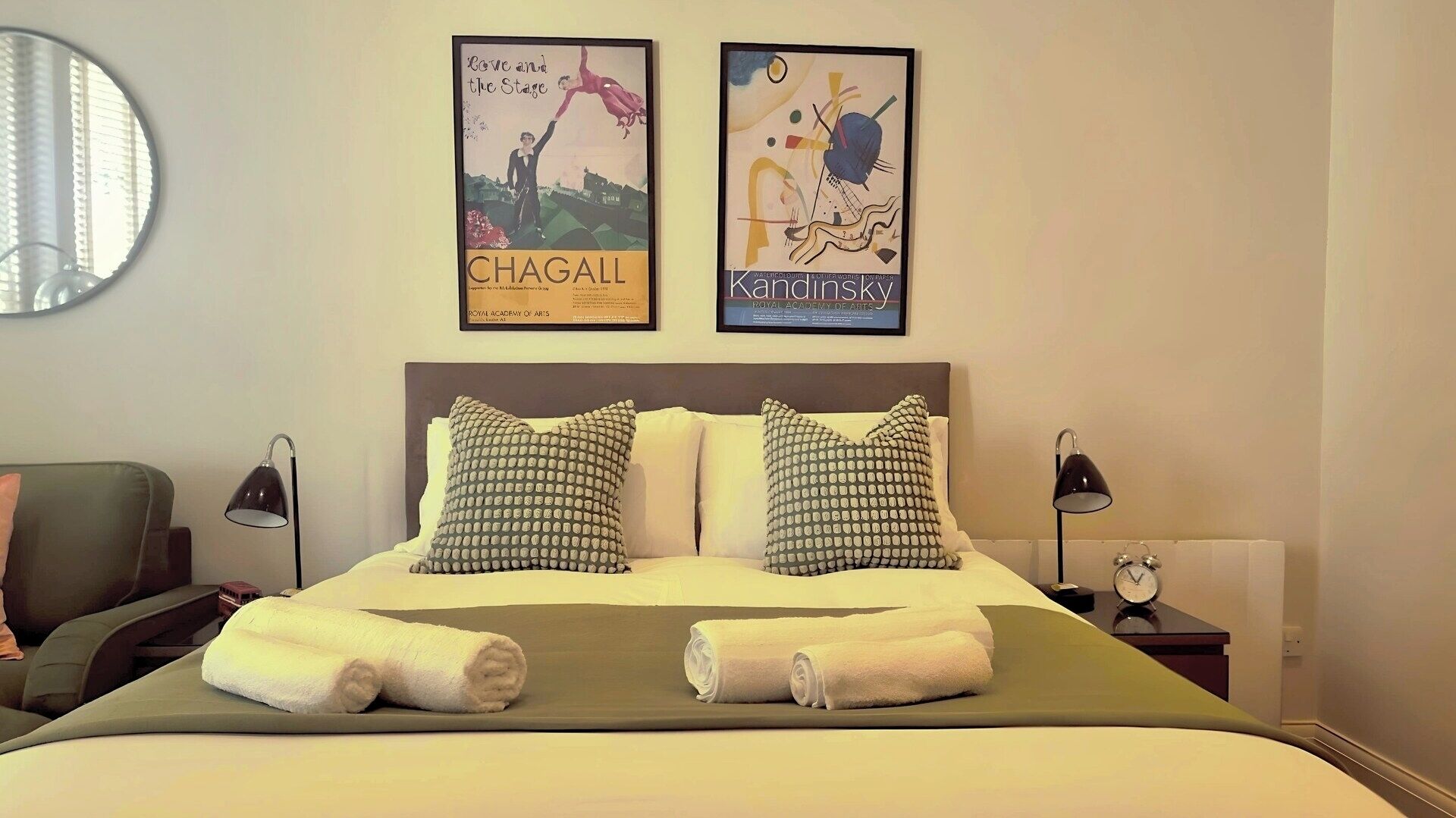 Een hotelkamer met een bed, twee kussens, een ronde spiegel en twee lijstjes met posters aan de muur.