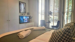 A hotel room with a bed, a desk with a chair, a TV on a stand, and a window with blinds. - Notting Hill - The One One One! -1- (London)