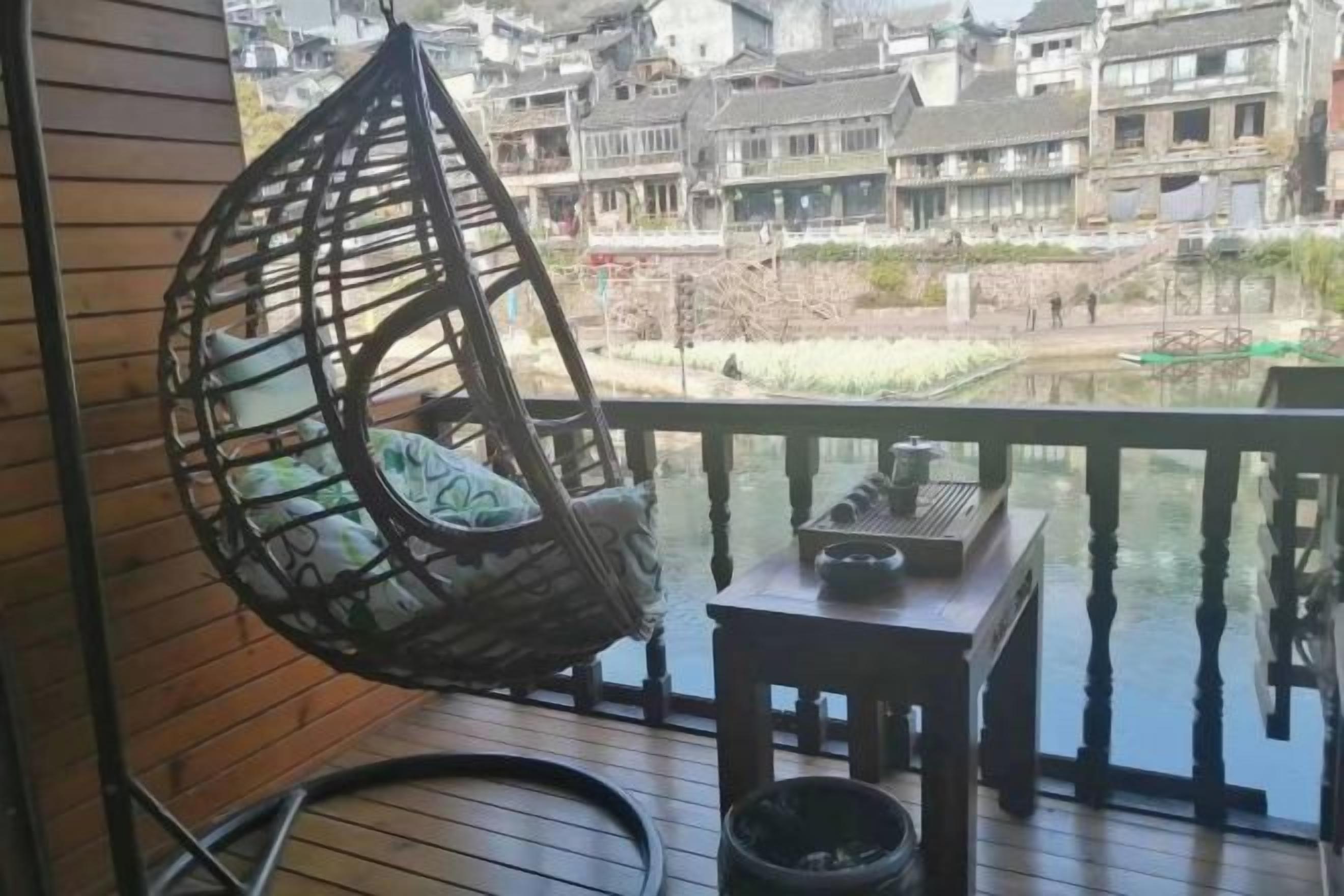 A wooden deck with a hanging chair, a small table, and a view of a river and buildings.