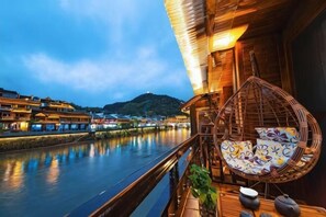 Ein Holzbalkon mit einem Korbstuhl, Blick auf einen Fluss und traditionelle Gebäude im Hintergrund.