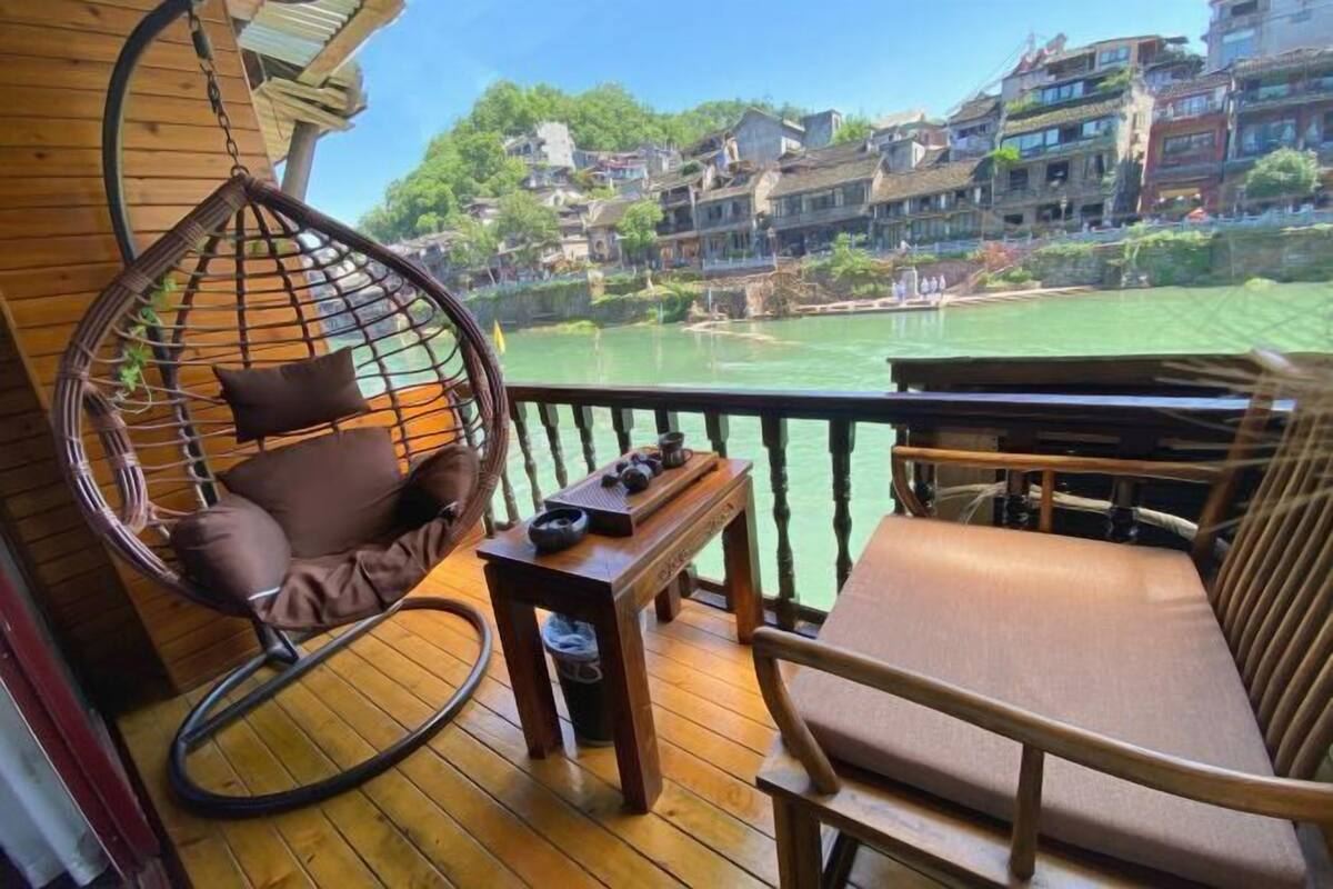 A wooden deck with a hanging chair and a cushioned bench, overlooking a river and traditional buildings.