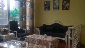 Um quarto com um sofá azul escuro, uma cadeira branca, uma mesa pequena com toalha florida e uma janela com cortinas.