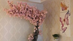 Quarto com uma árvore artificial decorativa com flores cor-de-rosa, uma parede com arte de borboletas e uma pequena planta em vaso sobre um suporte branco.