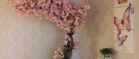 A room with a large artificial cherry blossom tree, a wall with a geometric pattern, and a small potted plant on a white stand.