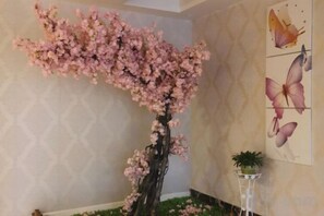 Una habitación con un gran árbol de cerezo artificial, una pared con un diseño geométrico y una planta pequeña en maceta sobre un pedestal blanco.