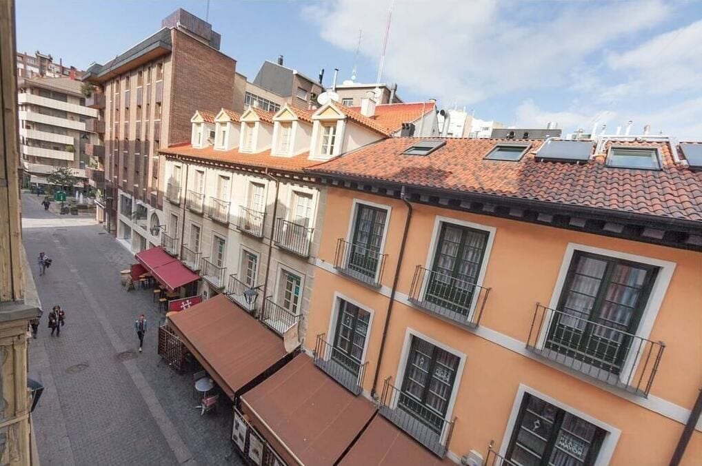 A street view of a building with multiple balconies, a red-tiled roof, and a pedestrian walkway.