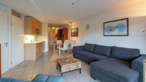 A modern living room with a grey sofa, a wooden coffee table, and a dining area with a white table and chairs. - Apartment in Callantsoog Near Beach Access (Callantsoog)