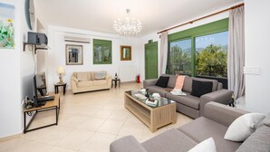 A modern living room with a sofa, a coffee table, and a TV. There is a green door and a window with a view of greenery. - Villa Poseidonia - Three Bedroom Villa, Sleeps 6 (Ionian Islands)
