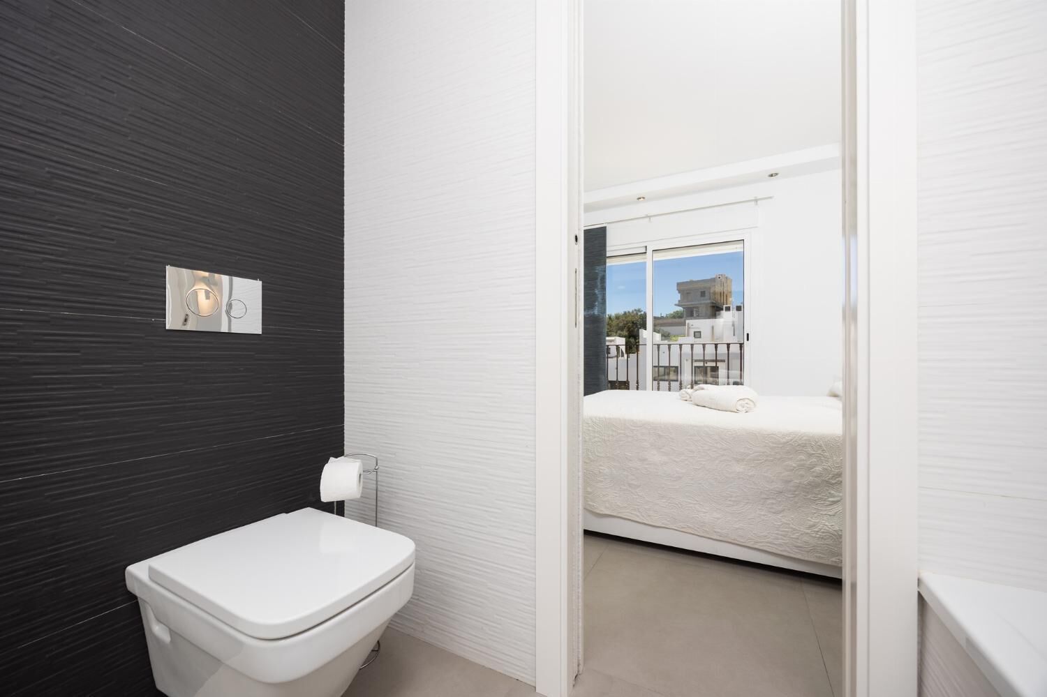 A modern bathroom with a toilet, a towel dispenser, and a window with a view of buildings.