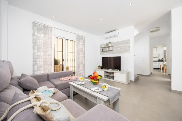 A modern living room with a grey sofa, a coffee table with fruit, a TV, and a kitchen in the background.