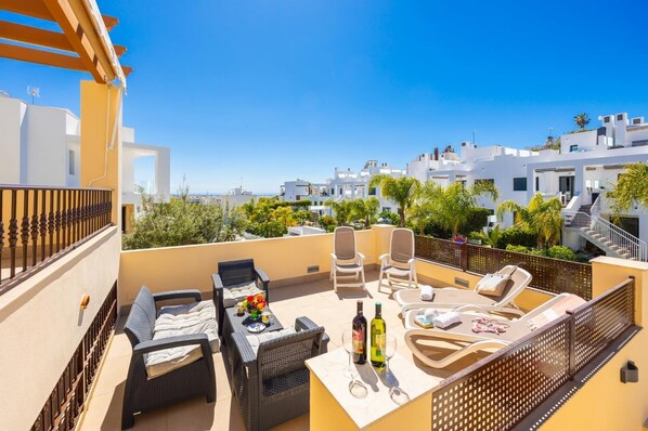 A rooftop terrace with outdoor furniture, a table with a bottle, and a view of white buildings and greenery.