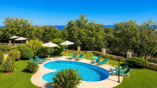 A swimming pool with lounge chairs and umbrellas, surrounded by greenery and a clear blue sky.