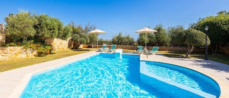 A rectangular swimming pool with clear blue water, surrounded by a tiled deck. There are lounge chairs and umbrellas along the poolside.