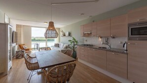 A modern kitchen with wooden cabinets, a dining area with wicker chairs, and a large window overlooking a scenic view. - Apartment in Groote Keeten Near the Beach (Callantsoog)