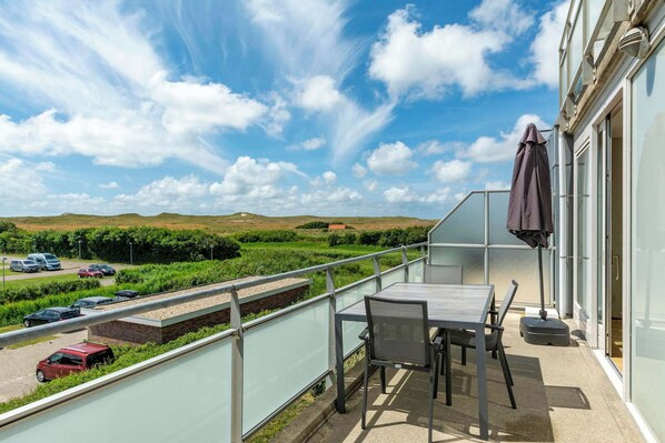 Ein Balkon mit Tisch und Stühlen, der auf einen Parkplatz und grüne Felder unter blauem Himmel blickt.