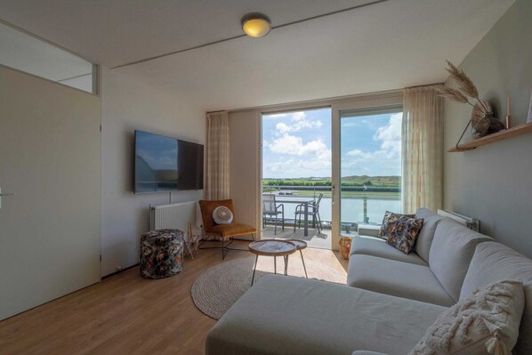 A modern living room with a sofa, a TV, a small table, and a view of a lake through large windows. - Apartment in Groote Keeten Near the Beach (Callantsoog)