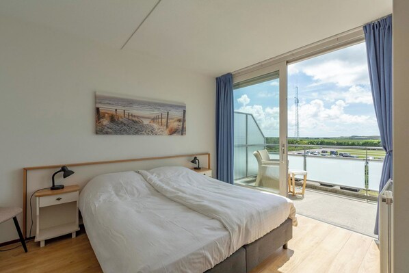 Une chambre à coucher avec un grand lit, une table de chevet, un bureau et une chaise. Il y a une fenêtre donnant sur un plan d’eau et un bâtiment.