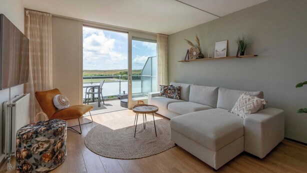 A modern living room with a sofa, a chair, a small table, and a view of the outdoors. - Apartment in Groote Keeten Near the Beach (Callantsoog)