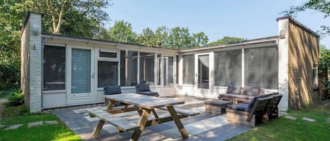 Eine überdachte Terrasse mit Holztisch und -stühlen, ein kleines Backsteinhaus und ein überdachter Bereich mit Moskitonetz.