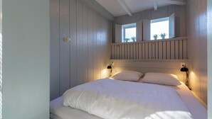 A bedroom with a bed, two bedside lamps, a window with plants, and a ceiling with exposed beams. - Duinland 110 Bungalow in Sint Maartenszee (Sint Maartensvlotbrug)