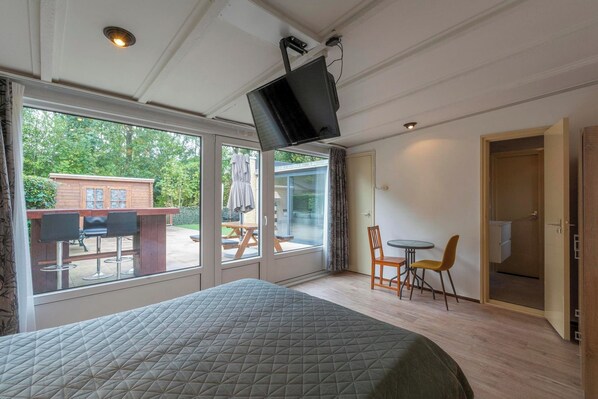 A bedroom with a bed, a TV mounted on the ceiling, a dining area with chairs and a table, and a sliding glass door leading to an outdoor patio.