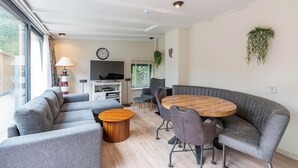 A modern living room with a curved grey sofa, a round wooden coffee table, and a grey armchair. There is a TV, a lamp, and a plant on the wall. - Bungalow Near Sint Maartenszee Beach (Sint Maartensvlotbrug)