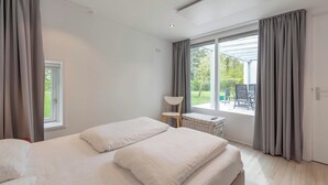 A modern bedroom with two beds, a window with curtains, a chair, and a wicker basket. - Duinland 146 for 4 Guests With Garden (Sint Maartensvlotbrug)