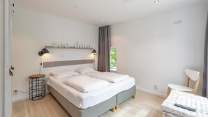 A modern bedroom with a double bed, bedside lamps, a small side table, a chair, and a window with curtains. - Duinland 146 for 4 Guests With Garden (Sint Maartensvlotbrug)