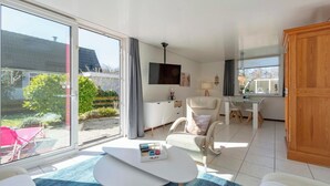 A modern living room with a large sliding glass door leading to a garden, a white sofa, a small white table, and a dining area with a white table and chairs. - Holiday Home St. Maartenszee Near Beach (Sint Maartensvlotbrug)