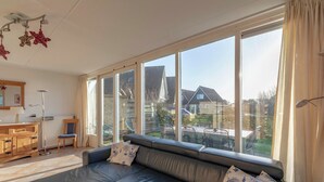 A modern living room with a black leather sofa, a wooden cabinet, and a large window offering a view of the backyard. - Callantsogervaart 3-8 Home Near Beach (Julianadorp)