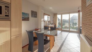 A modern kitchen with a built-in microwave, a dining area with a glass table and chairs, and a large sliding glass door leading to a balcony. - Callantsogervaart 3-8 Home Near Beach (Julianadorp)