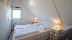 A small, white, attic-style bedroom with two beds, a window, and a nightstand with a lamp. - Callantsogervaart 3-8 Home Near Beach (Julianadorp)