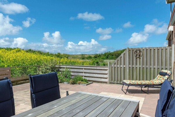 An outdoor patio with a table and chairs, a sun lounger, and a view of a garden with yellow flowers. - Apartment Groote Keeten Near Beach & Sauna (Callantsoog)