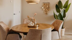 A dining area with a round wooden table, beige upholstered chairs, a hanging woven light fixture, a wall-mounted decorative piece, and a potted plant. - Apartment in Netherlands Near the Coast (De Cocksdorp)