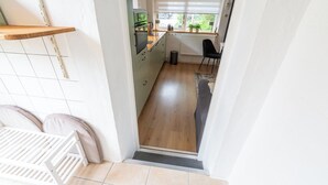Een smalle gang met een keukenhoek, een radiator, een raam met zonwering en een zwarte stoel.