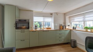 Een keuken met lichtgroene kastjes, een inbouwoven, een raam met zonwering, een radiator en een prullenbak.