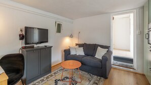 Een woonkamer met een grijze bank, een kleine oranje tafel, een zwarte stoel, een televisie op een grijze kast en een kamerbreed tapijt met een patroon.