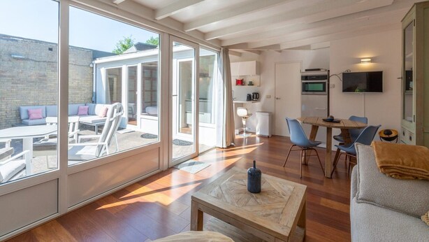 A spacious living area with large sliding glass doors leading to an outdoor seating area, a kitchen with a TV, and a dining table with chairs. - Duinland 122 Bungalow foe 2-people (Sint Maartensvlotbrug)