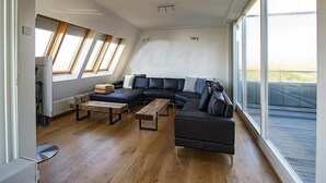 Ein modernes Wohnzimmer mit Ecksofa, Couchtisch und kleinem Hocker. Große Fenster sorgen für natürliches Licht und einen Blick ins Freie.